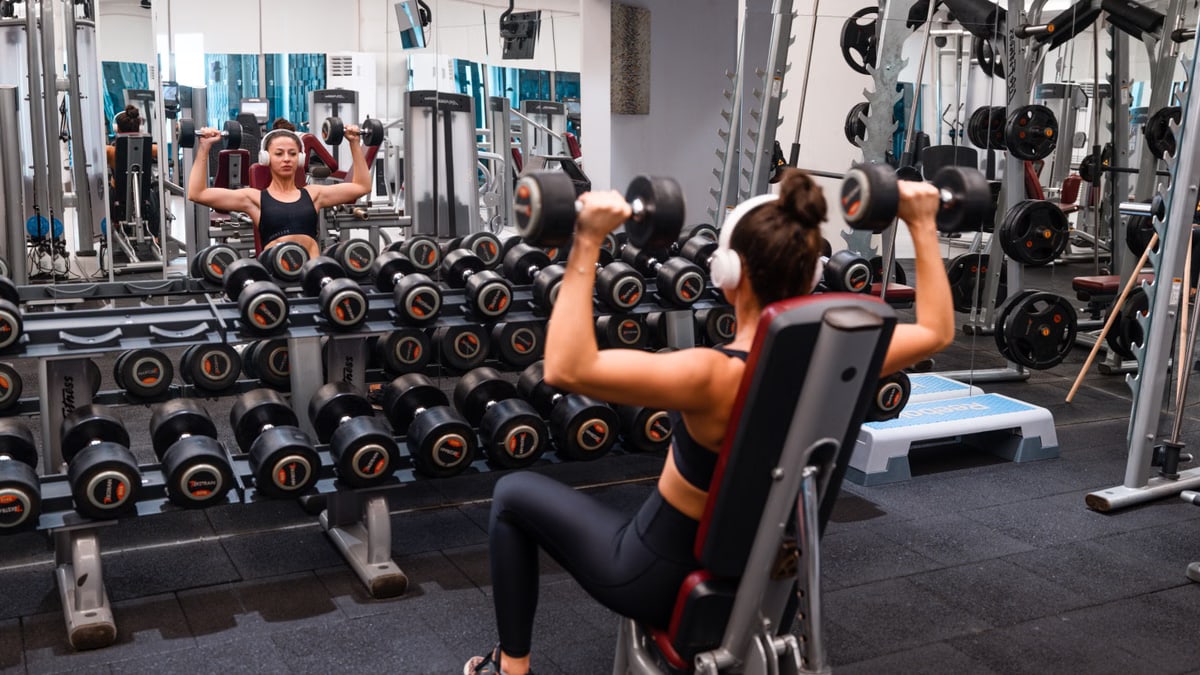 Woman exercising in the gym lifting dumbbells with headphones on for a strength-building workout.